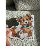 Kit de punto de cruz Andriana SANZ-77 Bookmark Friend con perrito marrón y blanco de ojos grandes, collar azul y diseño para marcapáginas sobre canvas plástico blanco.